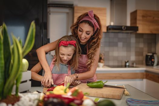 Cute little girl and her beautiful mom are cutting vegetables and smiling while cooking in kitchen at home.