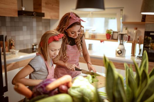 Cute little girl and her beautiful mom are cutting vegetables and smiling while cooking in kitchen at home.