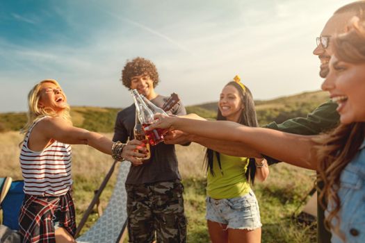 Happy young friends enjoy a sunny day in nature. They're clinking beer bottles, laughing and talking happy to be together.