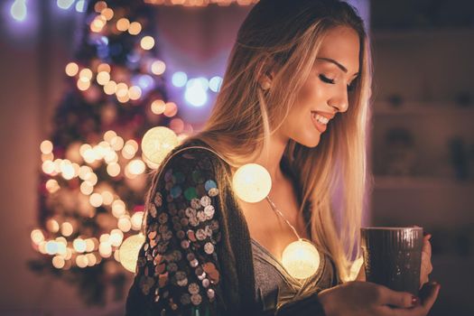 Cute young smiling woman holding cup of coffee, surrounded with Christmas bubble lights. 