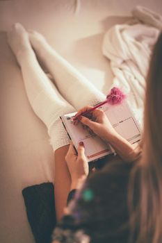 Cute young smiling woman sitting on the bed and making Christmas wish list in a her notes. 