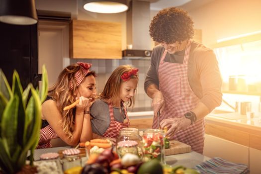 Cute little girl and her beautiful parents are cutting vegetables and smiling while making pickle in kitchen at home