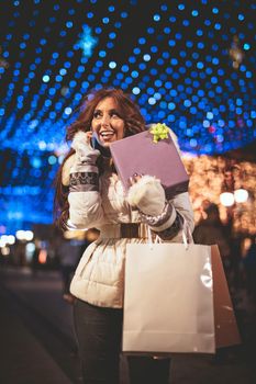 Cheerful young woman with colorful shopping bags using smartphone and having fun in the city street at Christmas time.