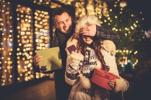 Young couple in the city centre with holiday's brights in background. Smiling man covering the eyes of happy surprised girlfriend in a Christmas night.