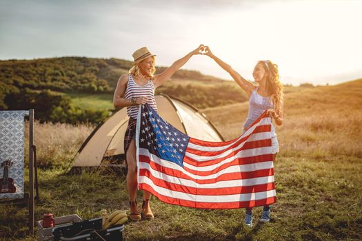 Happy young girl friends enjoys a sunny day in nature. They're holding an american flag in front a campsite tent. 