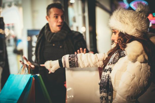 Young beautiful couple in the shopping in the city street at Christmas time. Cute girl praying her boyfriend that they are still in the shopping while they carrying many shopping bags.