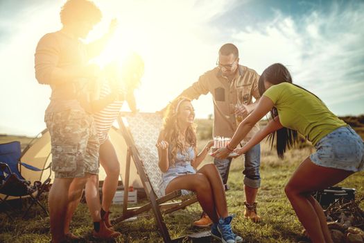 Young people have a good time in camp in nature. They're celebrating a birthday, laughing and greeting to their friend with birthday cake, happy to be together.