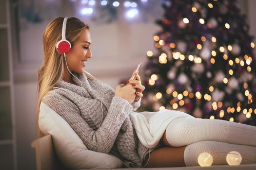 Cute young smiling woman listening music from smartphone during cozy Xmas holidays at home.