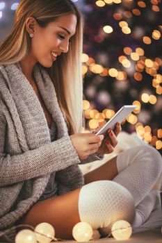 Cute young smiling woman using smarthphone and smiling during cozy Xmas holidays at home.
