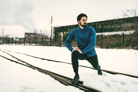 Active young man stretching and doing exercises in the public place among old railroad during the winter training outside in. Copy space. 