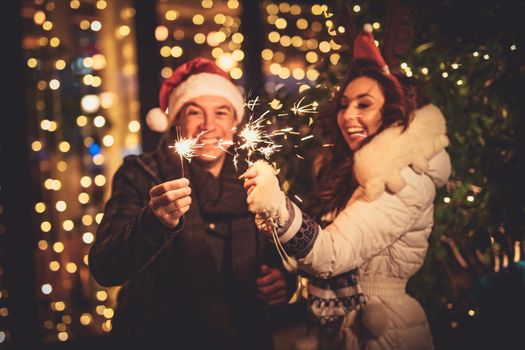 Cute cheerful couple with sparklers enjoying at the Christmas eve in the city street with a lot of holiday's lights.