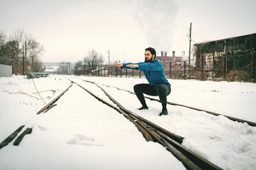 Active young man crouching and doing exercises in the public place among old railroad during the winter training outside in. Copy space.