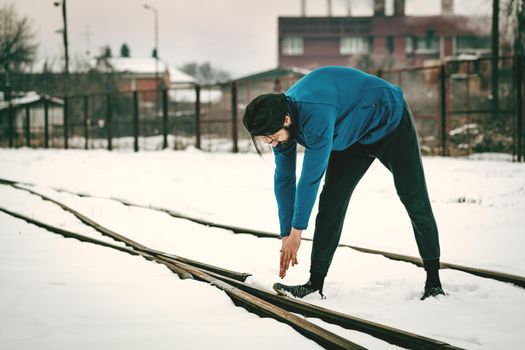 Active young  man stretching and doing exercises in the public place among old railroad during the winter training outside in. Copy space. 