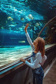 Young girl standing outstretched against aquarium glass fascinated by ocean world and touches the fishes in an oceanarium tunnel.