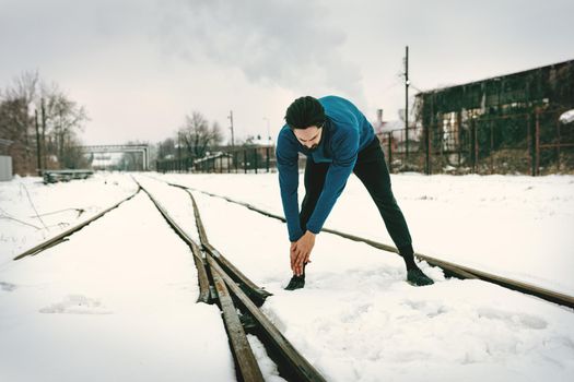 Active young man stretching and doing exercises in the public place among old railroad during the winter training outside in. Copy space. 