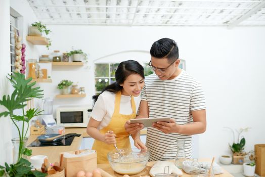 Young asian man and woman together cooking cake and bread with egg, looking menu from tablet in the flour happy relaxing in at home