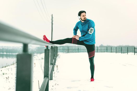 Active young man with headphones on his ears stretching and doing exercises in the public place during the winter training outside.