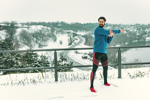 Active young man with headphones on his ears stretching and doing exercises in the public place during the winter training outside.