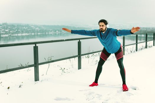 Active young man with headphones on his ears stretching and doing exercises in the public place during the winter training outside.