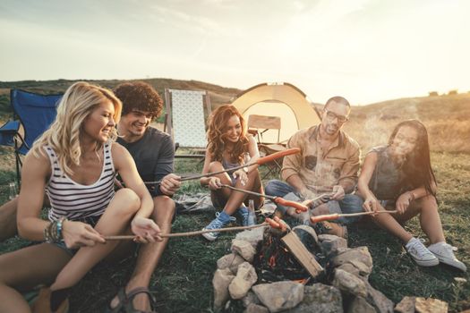 Happy young friends enjoy a sunny day at the mountain. They're laughing and grilling sausages on sticks over a campfire near tent.