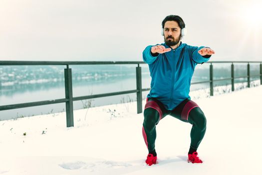 Active young man crouching and doing exercises in the public place among old railroad during the winter training outside beside the river. Copy space.