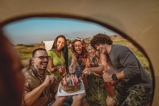 Young people have a good time in camp in nature. They're celebrating a birthday, laughing and greeting to their friend with birthday cake, happy to be together.