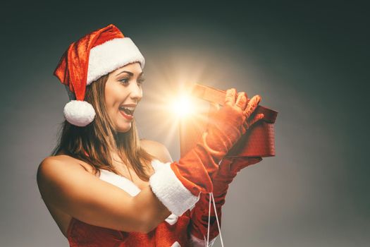 Beautiful young smiling woman in Santa Claus costume holding red present and having fun looking what is in it.