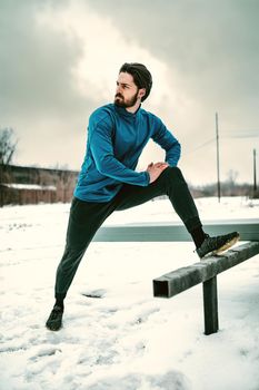 Active young man stretching and doing exercises in the public place during the winter training outside in while it snowing. Copy space.