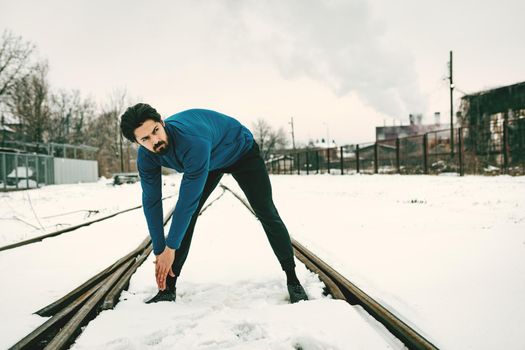 Active young man stretching and doing exercises in the public place among old railroad during the winter training outside in. Copy space. 