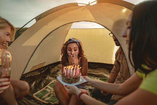 Young people have a good time in camp in nature. They're celebrating a birthday, laughing and greeting to their friend with birthday cake, happy to be together.