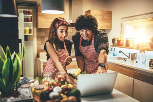 Young happy couple is enjoying and preparing healthy meal in their kitchen and reading recipes on the laptop.