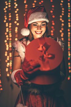 Beautiful young smiling woman in Santa Claus costume holding red present and having fun looking what is in it..