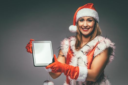 Beautiful young smiling woman in Santa Claus costume showing digital tablet and looking at camera. Selective focus.