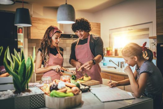 Happy parents and their daughter cooking together in the kitchen while little girl doing her homework on the kitchen table.