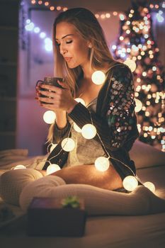 Cute young smiling woman sitting in the bed and holding cup of coffee, surrounded with Christmas bubble lights. 