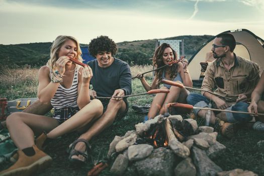 Happy young friends enjoy a sunny day at the mountain. They're laughing and roasting sausages on sticks over a campfire near tent.