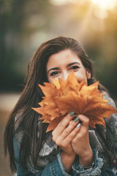 Cute young woman enjoying in sunny forest in autumn colors. She is holding many leaves and looking at camera behind leaves.