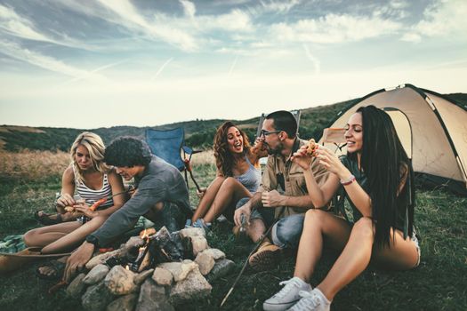 Happy young friends enjoy a nice day in nature. They're grilling sausages, eating, laughing and talking happy to be together.