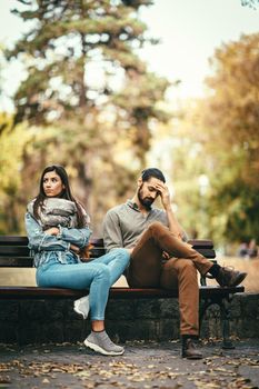 Young couple is sitting in the autumn city park on the bench, quarreling. They are angry and unhappy and not looking at each other.