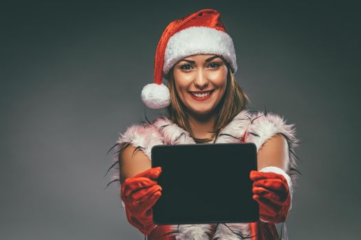 Beautiful young smiling woman in Santa Claus costume showing digital tablet and looking at camera. Selective focus.