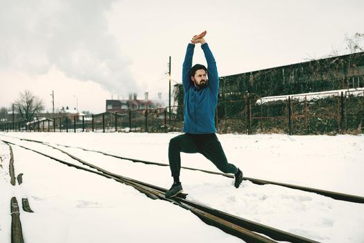 Active young man stretching and doing exercises in the public place among old railroad during the winter training outside in. Copy space. 