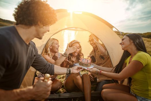 Young people have a good time in camp in nature. They're celebrating a birthday, laughing and greeting to their friend with birthday cake, happy to be together.