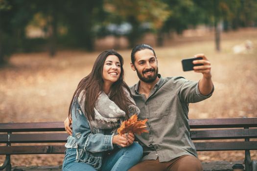 Beautiful smiling couple enjoying in sunny city park in autumn colors looking each other. They are sitting on the bench and having fun with smarthphone taking selfie.