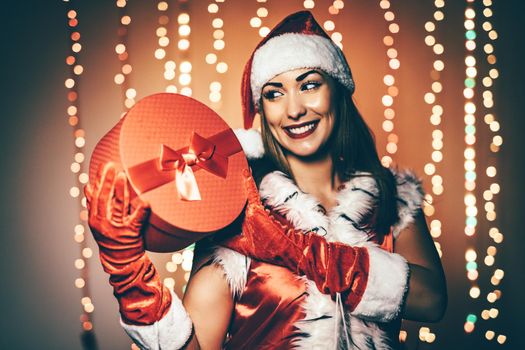 Beautiful young smiling woman in Santa Claus costume holding red present.