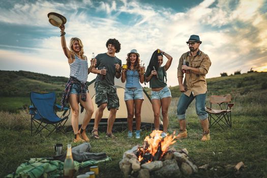 Happy young friends enjoy a sunny day at the mountain. They're laughing and preparing campfire near tent.