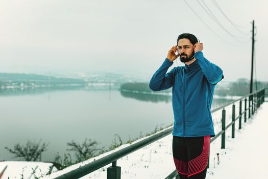 A male runner with headphones on his ears taking a break in the public place during the winter training outside beside the river. Copy space.