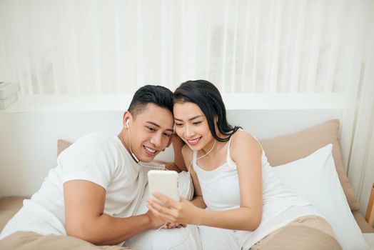 Indoor photo of loving couple lying on white sheets with smartphone.