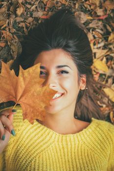 Cute young woman enjoying in sunny forest in autumn colors. She is lying down on the fall meadow looking at camera behind leaves.