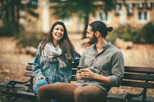 Beautiful smiling couple enjoying in sunny city park in autumn colors looking each other. They are sitting on the bench and having fun.
