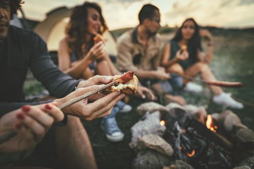 Happy young friends enjoy a nice day in nature. They're grilling sausages, eating, laughing and talking happy to be together.